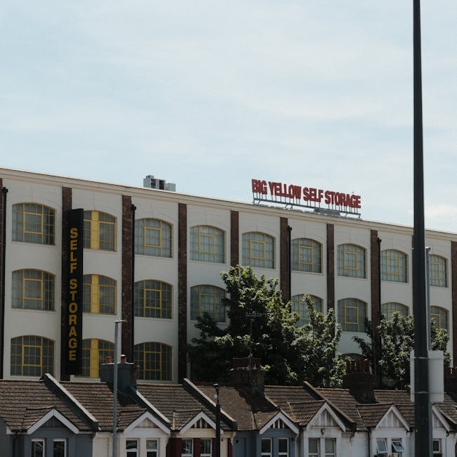 A multi-story modern residential building with a distinctive stepped design, featuring numerous rectangular windows and a dark brick exterior, situated near a body of water with a concrete retaining w
