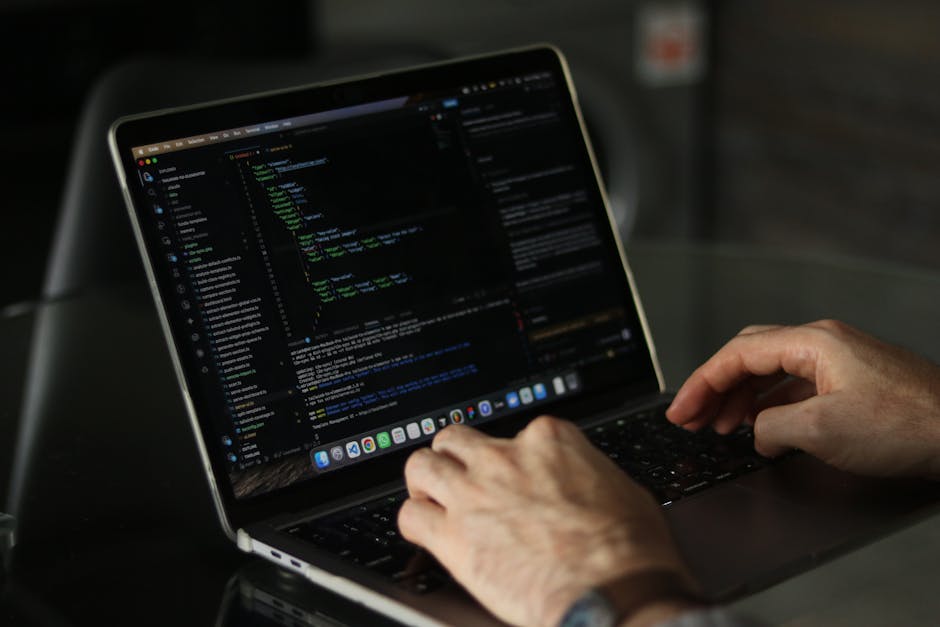 Close-up of a person's hands operating a laptop with a black keyboard, displaying a dark-themed code editor with lines of programming code in green, blue, and white text. The laptop is placed on a fla